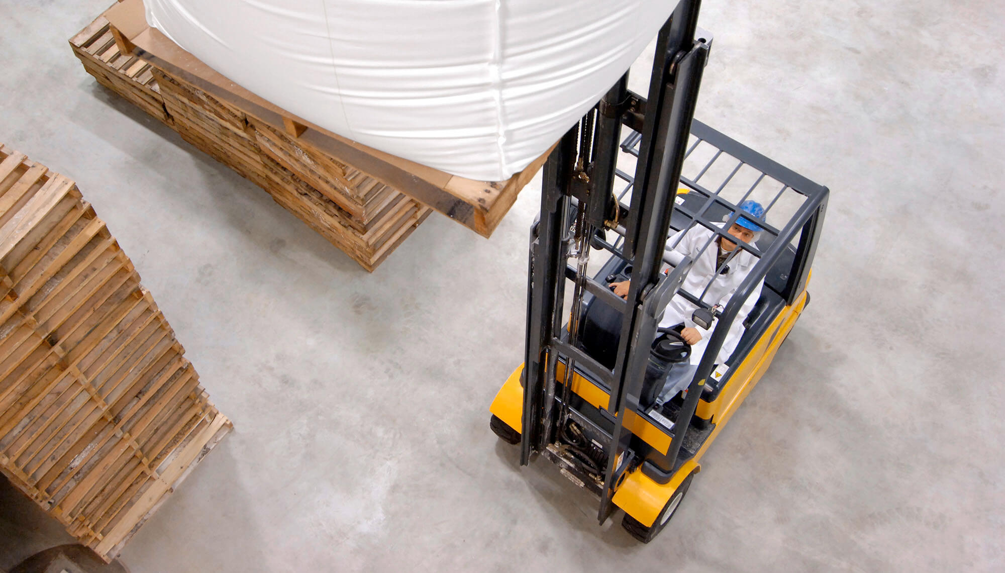 Aerial view of man in white jump suit and blue hard hat, driving a Battery-powered forklift truck in packaging warehouse