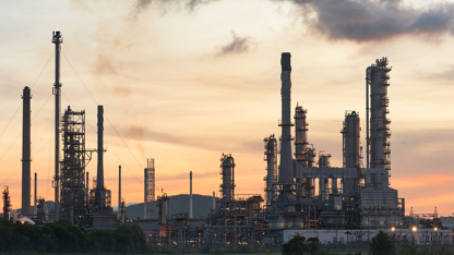 A refinery silhouetted against a sunset, with smoke billowing from its chimneys into the colorful sky.